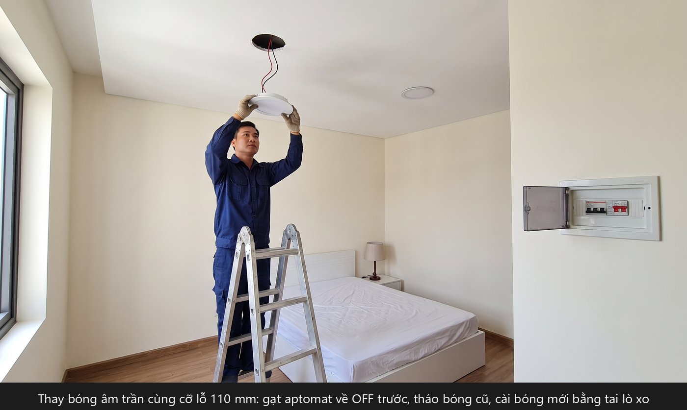 Thợ điện đứng thang nhôm thay đèn âm trần 12W lỗ khoét 110 mm trong phòng ngủ, tủ điện trên tường đã gạt aptomat về OFF